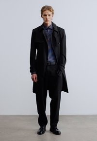 Μαύρο trench coat πάνω από πουκάμισο με μπλε μοτίβο, σε συνδυασμό με μαύρο παντελόνι και παπούτσια. Το ύφασμα φαίνεται λείο, με κομμένη εφαρμογή.