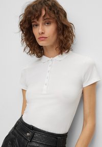 Biała koszulka polo z zapięciem na guziki, krótkim rękawem i subtelnym logo. Noszona z fakturowaną, czarną spódnicą o wysokim stanie.