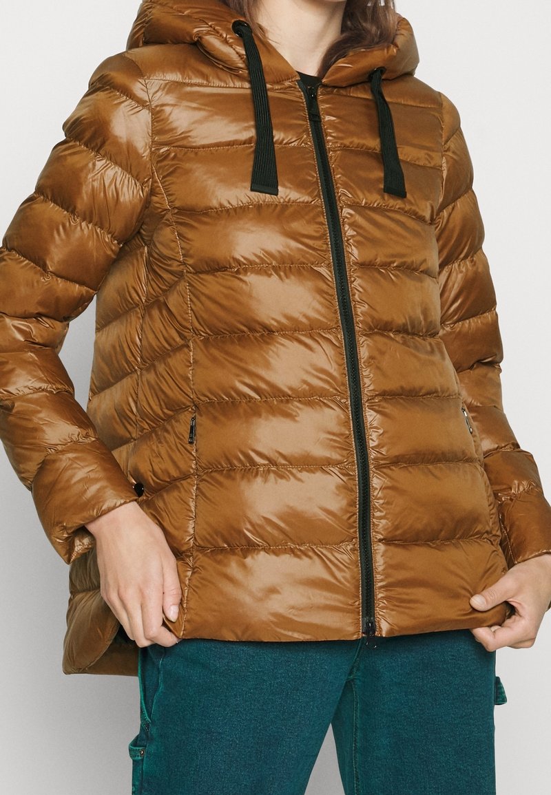 Personne portant une doudoune marron avec une fermeture éclair noire et des cordons de capuche, tenant l'ourlet de la veste, assortie à un pantalon bleu sarcelle.
