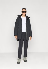 Chaqueta exterior negra con cremallera frontal, cuello alto y múltiples bolsillos. Combinada con una camisa blanca y pantalones oscuros, con zapatos grises.