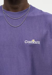 Κοντινό πλάνο ατόμου που φοράει μοβ μπλουζάκι Carhartt και δύο ασημένιες αλυσίδες κολιέ σε απλό φόντο.