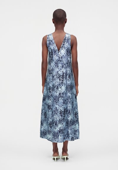 Personne aux cheveux courts portant une robe midi bleue à motifs sans manches avec un dos en V, debout devant un fond clair uni.