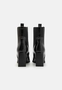 Paire de bottines en cuir noir avec talons carrés et languettes de tirage, vues de dos sur un fond blanc uni.