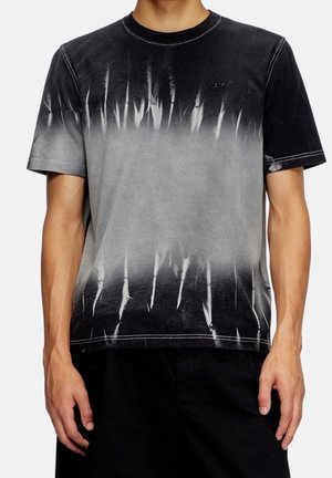 Hombre con camiseta de manga corta tie-dye en negro y gris con cuello redondo y pantalones negros sobre un fondo liso.