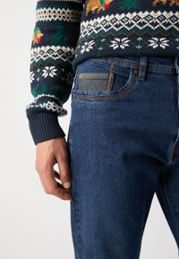 Marineblauwe trui met een multikleurig Fair Isle-patroon, met beren en bomen. Gedragen met blauwe denim jeans, die een textuurdetail in de zak tonen.