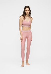 Λεπτή γυναίκα φοράει ροζ αθλητικό σουτιέν και κολάν με το ίδιο σχέδιο στη μέση, στέκεται ξυπόλυτη σε απλό λευκό φόντο.