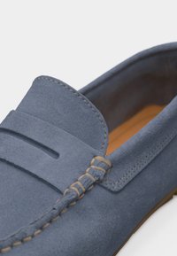 Μπλε μοκασίνι από σουέντ με μπεζ ραφές και λεπτομέρεια με λουράκι στο πάνω μέρος, με ανοιχτό καφέ εσωτερική επένδυση και σόλα.