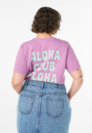 Lyhyttukkainen kiharatukkainen henkilö pukeutuu violettiin t-paitaan, jossa lukee "ALOHA CLUB ALOHA", ja korkeavyötäröisiin sinisiin farkkuihin, selin kameraan.