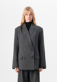 Blonde Frau trägt ein übergroßes, dunkelgraues, doppelt geknöpftes Blazer und passende weit geschnittene Hosen, steht vor einem schlichten weißen Hintergrund.
