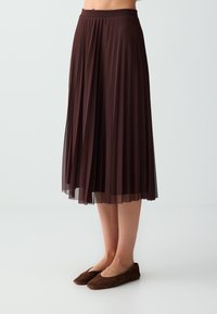 Jupe midi plissée marron foncé portée avec des chaussures plates marron foncé assorties, montrée de la taille aux pieds sur fond uni.