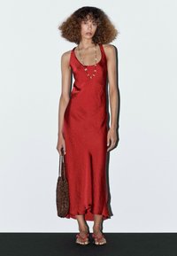 Červené saténové šaty s volným lemem, doplněné perlovým náhrdelníkem a hnědou tkanou taškou. Modelka má na sobě červené sandály na ramínka.
