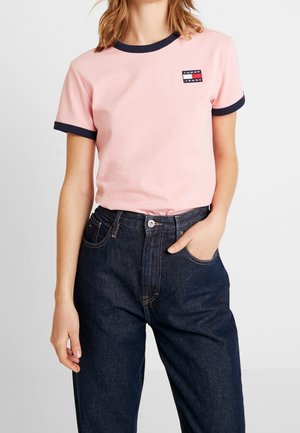 Kvinde iført en lyserød kortærmet skjorte med navy kant og Tommy Jeans logo, kombineret med mørkeblå jeans med høj talje.