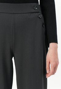 Primo piano di pantaloni neri a vita alta con quattro bottoni neri sul lato, indossati con un top nero a coste a maniche lunghe e una mano rilassata.