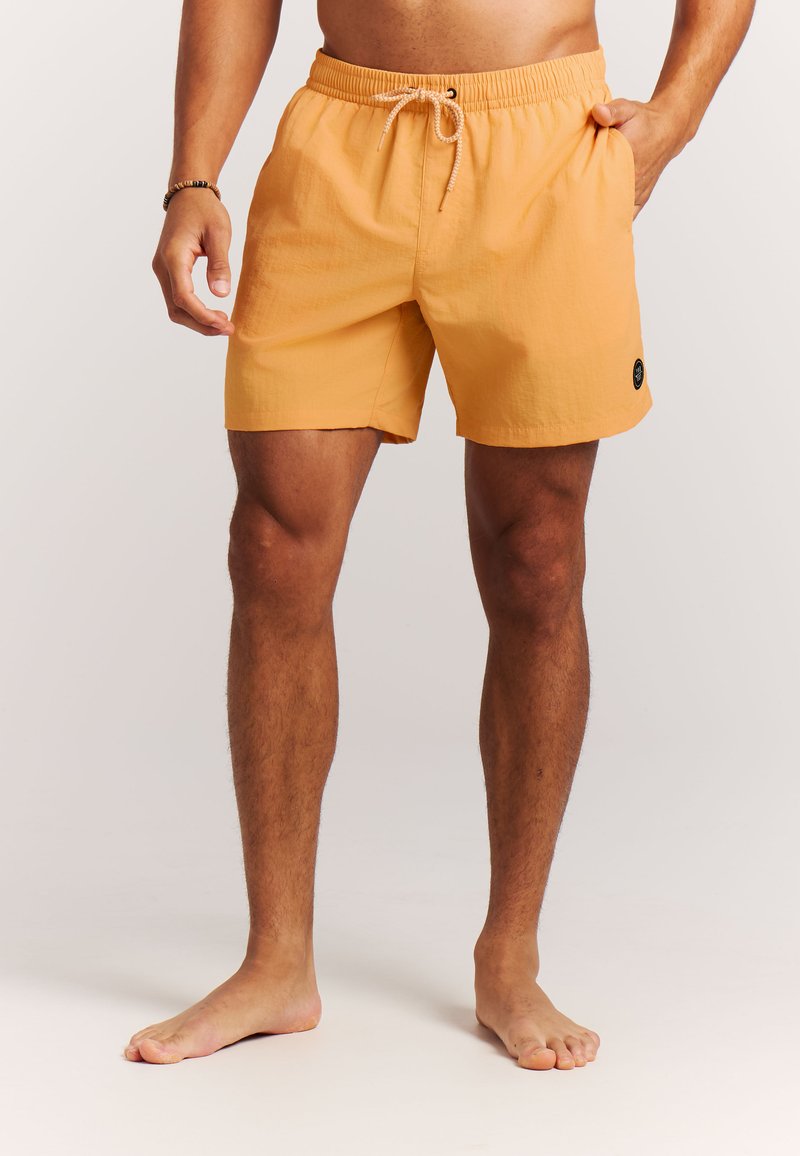 Uomo in costume da bagno arancione con coulisse in vita, in piedi a piedi nudi su sfondo chiaro.