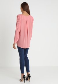Blouse rose à manches longues avec une texture lisse, pli central au dos, ourlet incurvé, portée avec un jean skinny foncé et des talons hauts noirs.