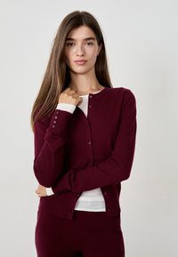 Jeune femme aux longs cheveux bruns portant un cardigan bordeaux boutonné sur un top blanc, se tenant contre un fond clair et uni.
