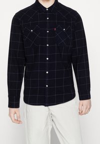 Marinblå rutig skjorta med knappar i flanell, med vita rutmönster, två bröstfickor och silverfärgade tryckknappar. Långa ärmar, avslappnad passform.