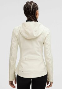 Light cream athletic jacket with a fitted design, hood, and textured fabric. Features a zippered pocket and breathable details at the back.