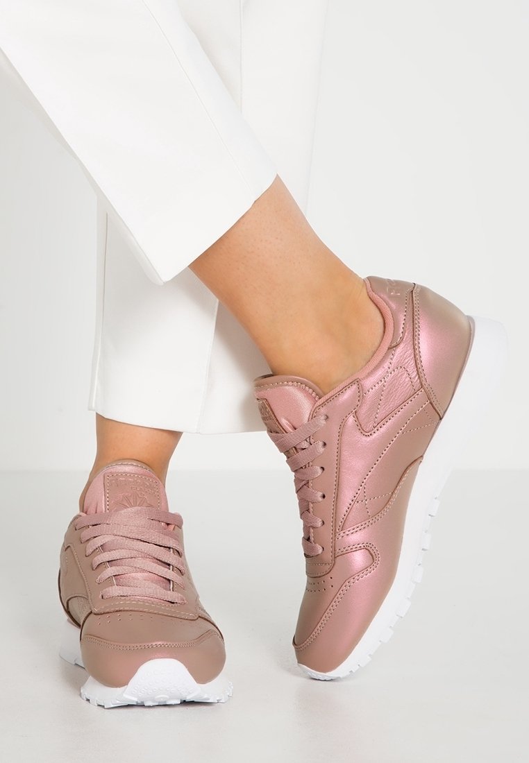 Baskets de sport en or rose avec une finition lisse, des accents texturés, un design à lacets et des semelles en caoutchouc blanches. Portées avec un pantalon blanc.