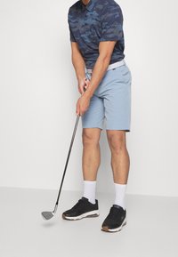 Niebieska koszulka polo w kamuflaż, z krótkimi rękawami, jasnoniebieskie spodenki i czarne buty sportowe; zawodnik trzyma kij golfowy w gotowości do uderzenia.