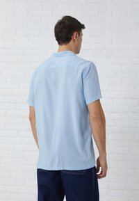 Chemise à manches courtes en tissu bleu clair avec une texture lisse, col classique et coupe droite. Design simple, sans motifs ni accents.