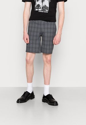 Pantalones cortos de cuadros grises con líneas azules contrastantes, combinados con una camiseta gráfica negra, calcetines blancos y zapatos negros. Diseño de corte recto.