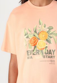 Camiseta de algodón de color melocotón con mangas cortas que presenta un gráfico floral de naranjas y hojas verdes, y el texto "CADA DÍA ES UN NUEVO COMIENZO."