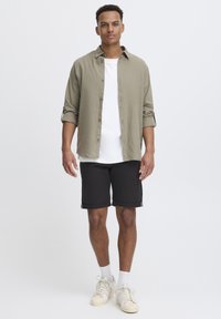 Chemise vert olive clair, T-shirt blanc et shorts noirs. La chemise a des manches retroussées ; les shorts sont à ourlets repliés. Des baskets beiges complètent la tenue.