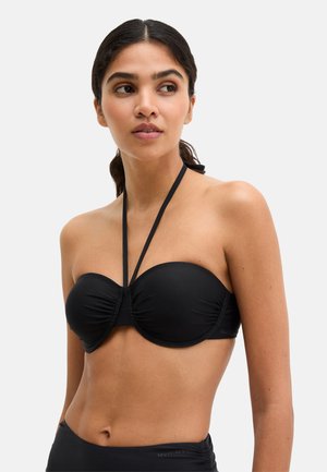 Frau mit dunklen Haaren in einem schwarzen trägerlosen Bikini-Top mit Neckholder, die leicht nach links blickt vor einfachem Hintergrund.