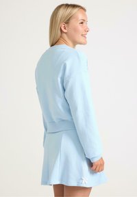 Felpa azzurra chiara con polsini e orlo a costine, abbinata a una gonna pantalone coordinata con dettaglio di nodo laterale. Tessuto liscio e elastico.