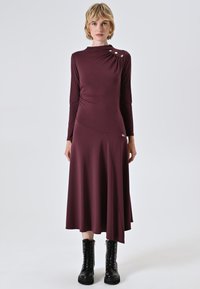 Langärmliges, burgunderrotes Kleid mit hohem Ausschnitt, asymmetrischem Saum und seitlicher Raffung, akzentuiert durch drei goldene Knöpfe an der Schulter.