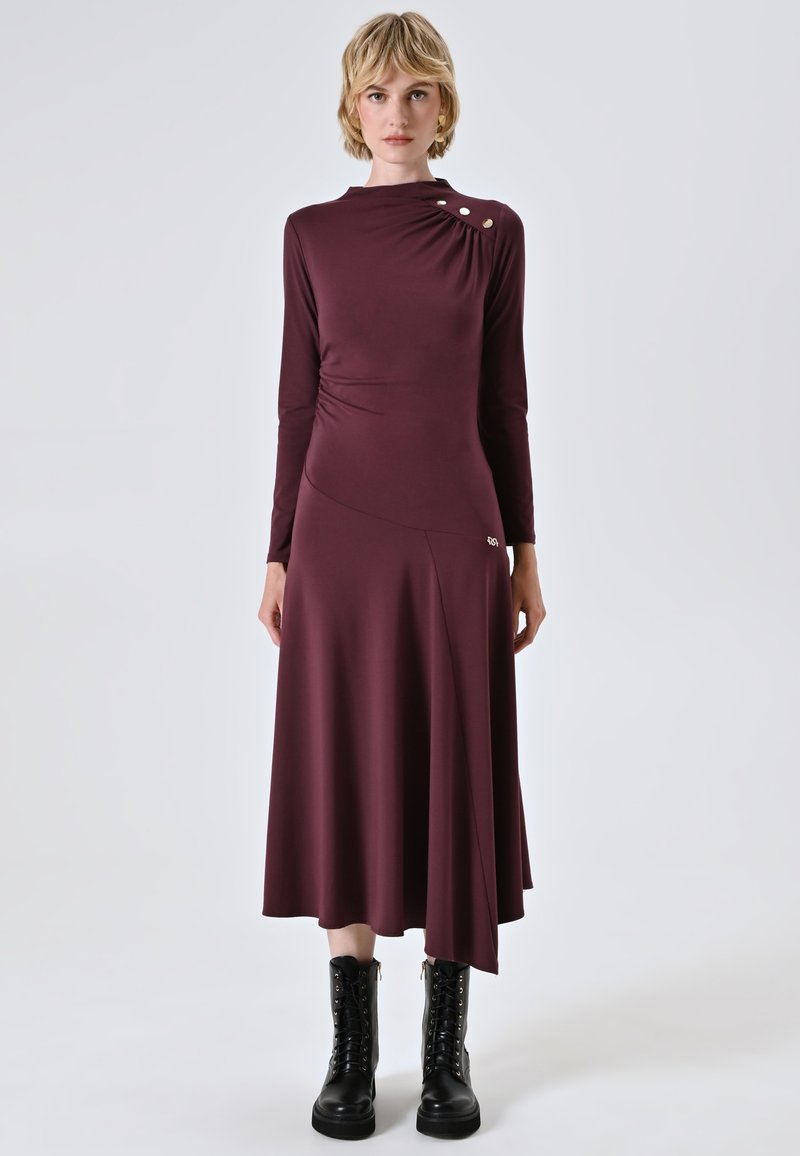 Langärmliges, burgunderrotes Kleid mit hohem Ausschnitt, asymmetrischem Saum und seitlicher Raffung, akzentuiert durch drei goldene Knöpfe an der Schulter.