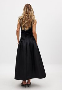Vestido maxi de satén negro con cintura fruncida, diseño sin mangas y bolsillos laterales, combinado con sandalias negras de tiras.