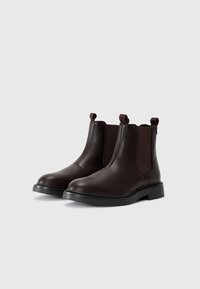 Botas Chelsea de cuero marrón con paneles laterales elásticos, punta redondeada y acabado texturizado. Presentan lengüetas de tira y una suela negra contrastante.