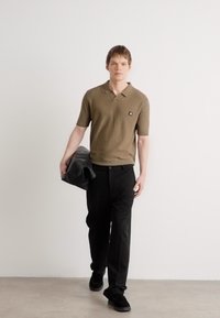 Koszulka polo z krótkim rękawem w oliwkowym kolorze, czarne spodnie i czarne buty. Model nosi czarną skórzaną torbę, prezentującą czyste linie i prosty design.