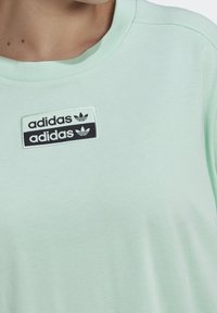 Světle mátově zelené bavlněné tričko s kulatým výstřihem a černobílou nášivkou loga Adidas na přední straně. Hladká textura.