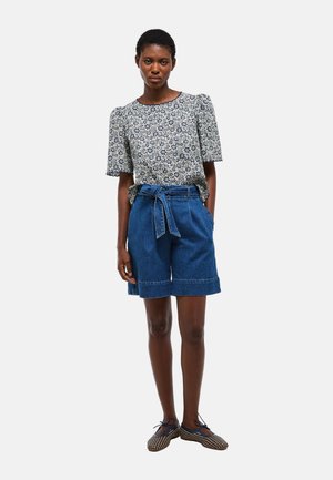 Femme debout portant un chemisier fleuri à manches courtes, un short en jean taille haute avec ceinture nouée, et des chaussures plates à motifs sur fond uni.