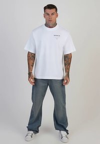 White cotton t-shirt with short sleeves, crew neckline, and black logo on the chest, paired with blue denim pants and white sneakers.