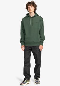 Haut à capuche vert avec cordon de serrage, poignets et ourlet côtelés ; associé à un jean foncé et des baskets multicolores. Le modèle se tient droit.