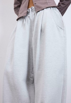 Henkilö pukeutunut vaaleanharmaisiin, löysälahkeisiin collegehousuihin, joissa on joustava vyötärönauha ja sivutaskut, yhdistettynä ruskeaan raidalliseen paitaan.