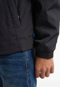 Jachetă neagră cu o textură netedă, manșete elastice și un mic eticheta albastră. Asortată cu blugi din denim albastru, cu cusături standard.