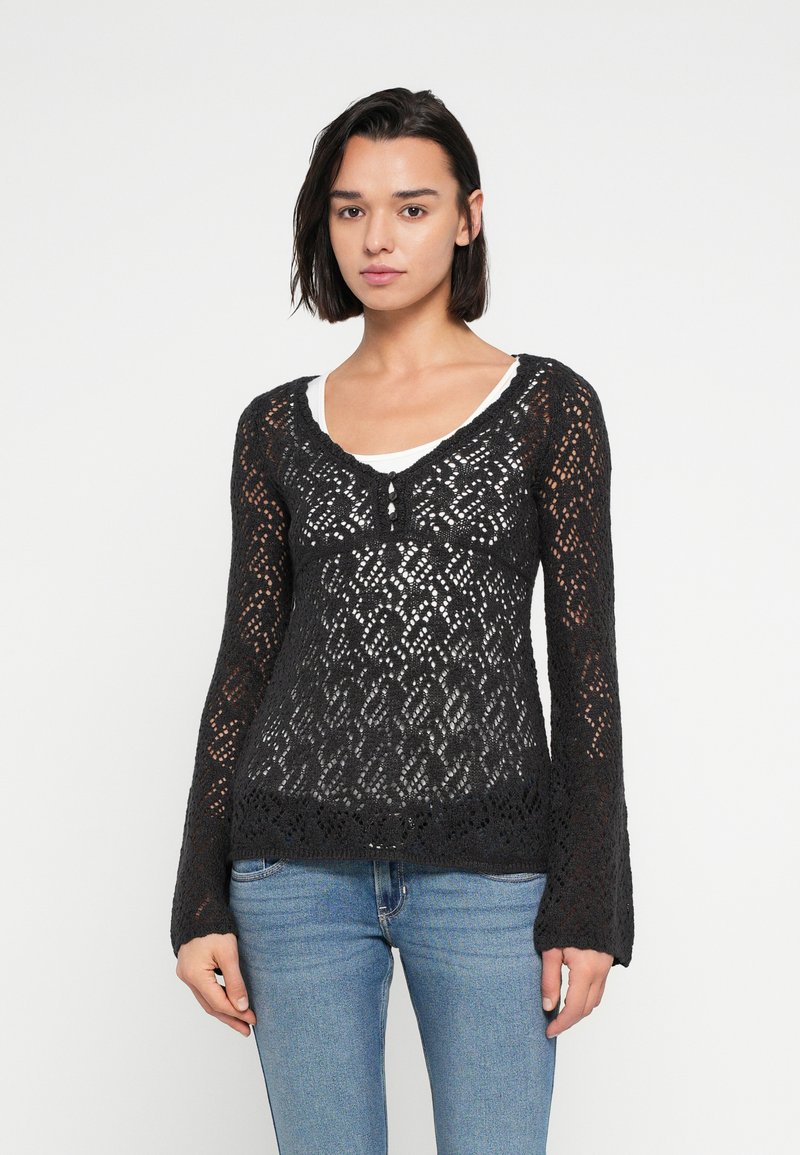 Μαύρο μακρυμάνικο τοπ από δαντέλα με πλέξη, με V-λαιμόκοψη, λεπτομέρεια με κουμπιά στο μπροστινό μέρος και εφαρμοστή γραμμή. Φορεμένο πάνω από ένα λευκό μπλουζάκι.