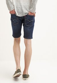 Tummansiniset farkkushortsit, joissa on klassinen viiden taskun muotoilu, käännetyt lahkeet ja huomaamattomat takataskun ompeluaksentit. Mukauttavat kengät esitelty.