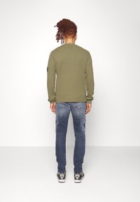 Olivgrön långärmad skjorta med rund halsringning, kombinerad med ljusblå jeans och grå sportskor med gröna accenter.