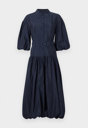 Frau steht, trägt ein marineblaues, geknöpftes Kleid mit Puffärmeln, einem Taillengürtel und schwarzen spitzen Absatzschuhen.