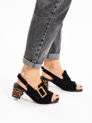 Feet wearing black open-toe slingback sandals with leopard print block heels and large buckles, paired with rolled-up gray jeans.