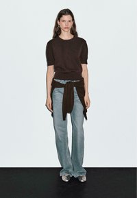 Top en tricot marron à manches courtes, jean en denim bleu clair évasé avec un ourlet brut, et chaussures argentées à enfiler. Pull marron noué à la taille.