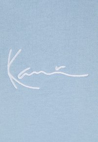 Svijetloplavi tekstil s bijelim vezenim potpisom. Tekstura je glatka s finim pletenim uzorkom.