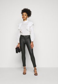 Blusa branca com pregas e detalhe em renda, combinada com calças justas verde-escuras e saltos altos pretos com tiras. A modelo segura uma carteira preta.