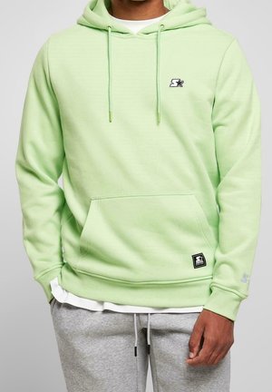 Person trägt hellgrünen Hoodie mit Fronttasche und kleinem Stern-Logo, kombiniert mit grauer Jogginghose und weißem Unterhemd.